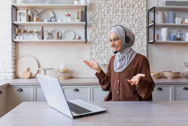 Радостная женщина в хиджабе работает из дома удаленно, мусульманка разговаривает по видеосвязи с помощью гарнитуры и ноутбука, деловая женщина работает удаленно на дому. - Фото, изображение