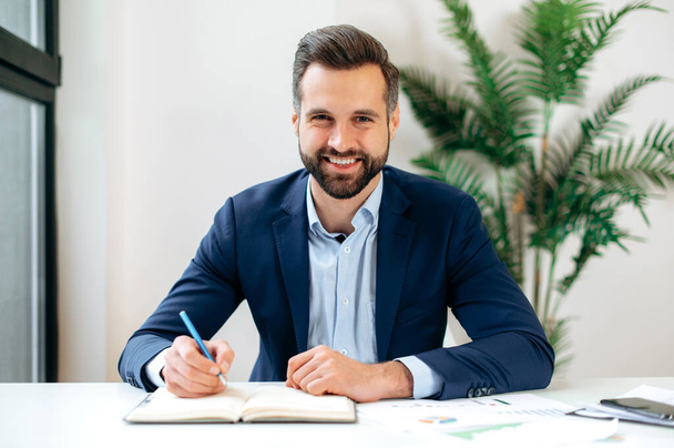 Positive attractive caucasian professional business man, mentor, looking in a webcam during video conference, listens lecture, leading webinar, record online training, takes notes, smiling friendly - Foto, Bild