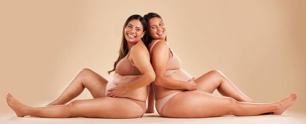 Pregnant woman friends, studio and underwear with smile, sitting and together for support by backdrop. Happy women, pregnancy and solidarity for wellness, care and holding healthy stomach by backdrop. - Fotó, kép