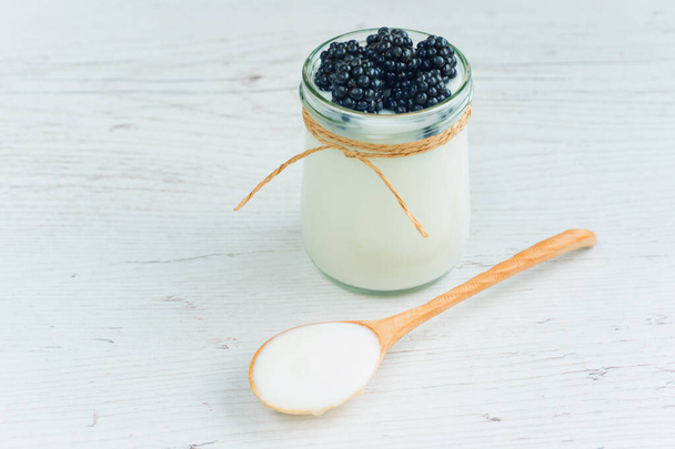 Yogurt in small jar with fresh berries. Blackberries in yogurt. Yogurt is in jar on the wooden table. Wooden spoon full of yogurt lies on the table - Zdjęcie, obraz