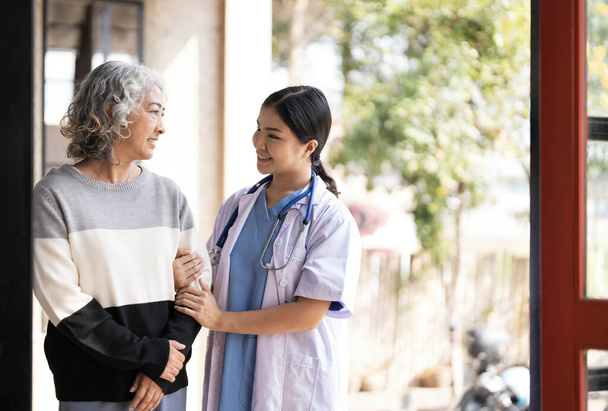 Jeune aidante aidant une femme âgée à marcher. Infirmière aidant sa patiente âgée à la maison de retraite. Femme âgée avec bâton de marche aidée par une infirmière à la maison... - Photo, image