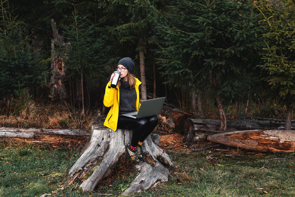 Woman wearing glasses and yellow jacket working on laptop outdoors surrounded by beautiful nature, forest and mountains. Workation concept. Freelancer work - Foto, Imagem