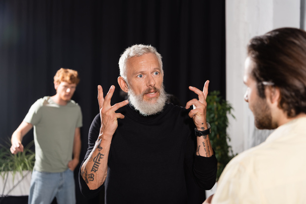tattooed and bearded art director gesturing while talking to blurred actor in theater. Translation of tattoo: "peace" - Fotografie, Obrázek