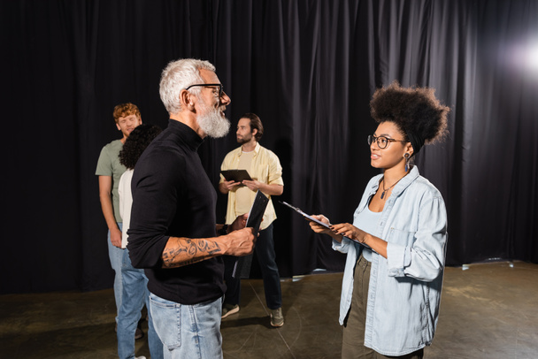 tattooed grey haired screenwriter holding scenario near african american woman with clipboard and actors in theater - Photo, image