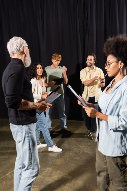 multiethnic actors holding clipboard with scenarios near grey haired screenwriter in theater - Photo, image