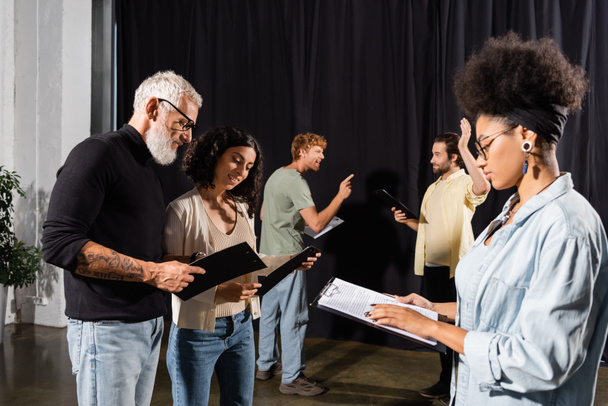 african american woman reading screenplay near bearded art director and troupe rehearsing in theater - Foto, Imagen