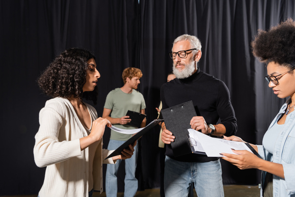 bearded acting skills teacher looking at interracial students holding clipboards with screenplays - Фото, изображение