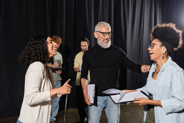 cheerful african american woman holding screenplay and laughing near bearded producer and multiethnic actors in theater - Photo, image