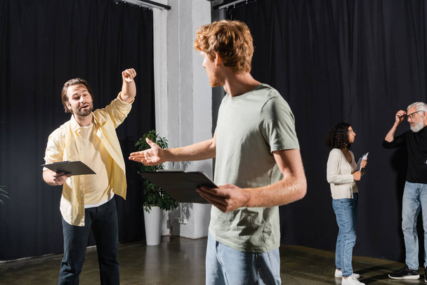 redhead and bearded actors holding clipboards with screenplays while rehearsing in theater  - Foto, afbeelding