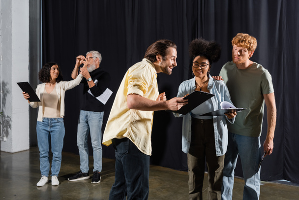 smiling man showing screenplay to interracial actors near art director and multiracial woman rehearsing on background - Photo, Image