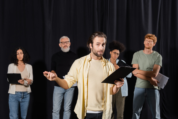 brunette bearded man holding clipboard and looking at camera while rehearsing near multicultural actors and art director in theater - Photo, Image