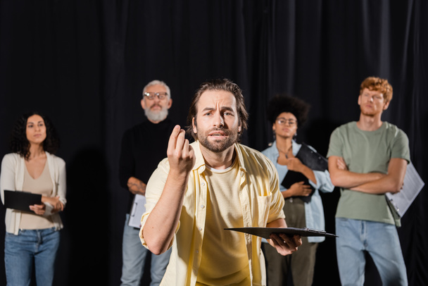 emotional man with clipboard looking at camera and gesturing during rehearsal near multiethnic actors and producer on blurred background - Photo, image