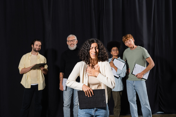 smiling multiracial woman holding clipboard and smiling near interracial actors and art director on blurred background - Photo, image