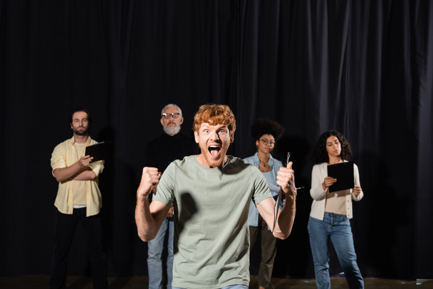 emotional redhead man showing thumb up and screaming near multiethnic students and teacher of acting skills on background - Foto, immagini