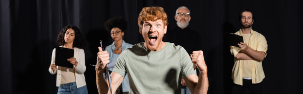 excited redhead man shouting and showing like gesture near producer and multiethnic actors on blurred background, banner - Photo, image