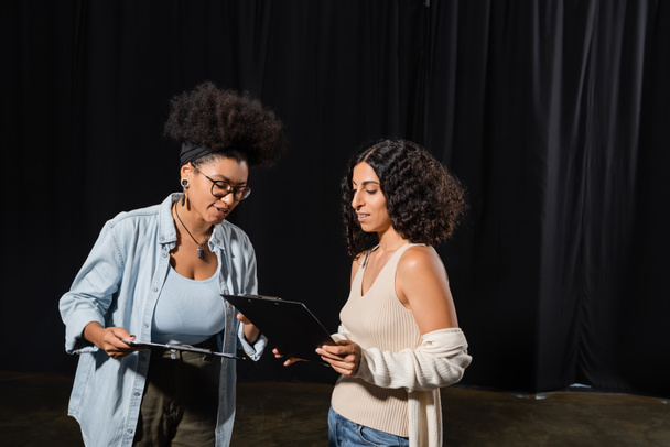 young interracial actresses looking at clipboards with scenarios in theater - Фото, изображение