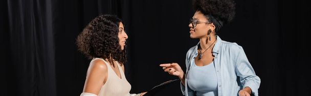 smiling african american woman in eyeglasses pointing with finger while talking to multiracial actress in theater, banner - Fotó, kép