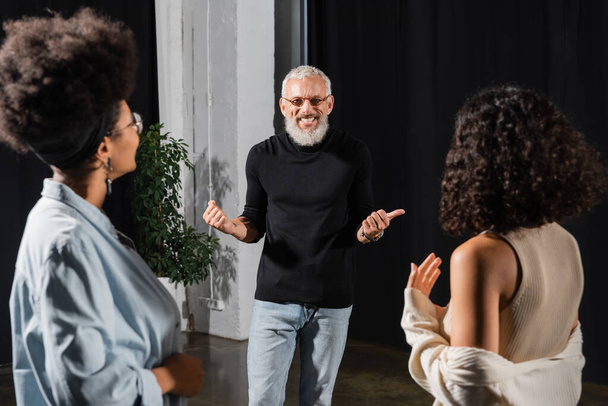 cheerful grey haired man pointing with thumb near multiethnic actresses in acting skills school - Fotoğraf, Görsel