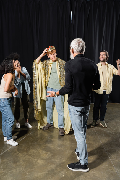 redhead man adjusting king crown near cheerful interracial actors and grey haired producer gesturing on foreground - Foto, imagen