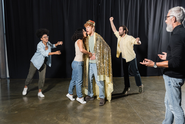 bearded producer gesturing near interracial students rehearsing scene of king assassination in theater - Φωτογραφία, εικόνα