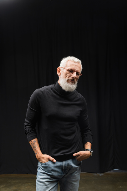 mature tattooed actor in eyeglasses and black turtleneck standing with hands in pockets of jeans in theater - Fotografie, Obrázek