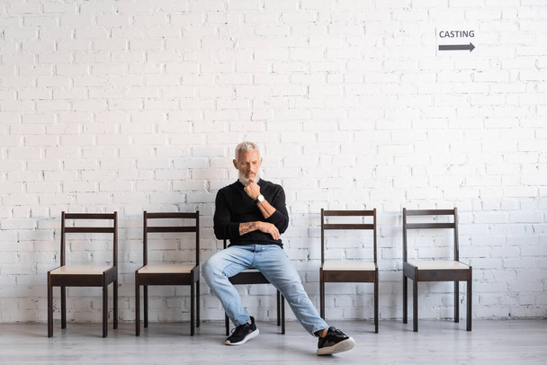 повнометражний бородатий чоловік сидить у залі біля білої стіни і чекає на кастинг. Переклад татуювання: om, shanti, peace
 - Фото, зображення