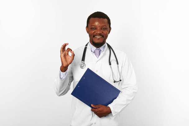 Smiling black bearded doctor man in white robe with stethoscope holds medical clipboard and shows OK gesture, isolated on white background. Happy black african american physician therapist portrait - Foto, afbeelding