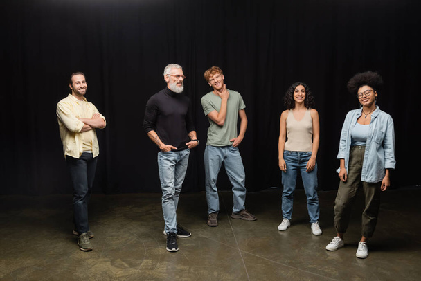 smiling bearded art director standing with hands in pockets near young multiethnic actors on stage in theater - Fotografie, Obrázek