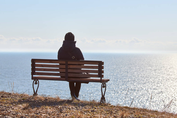 Single girl in a black jacket and hat sitting on bench at cliff at front of sea, peaceful and quiet place for thinking alone, loneliness and loss of loved one concept. Pacifying view of marine horizon - Foto, Imagem