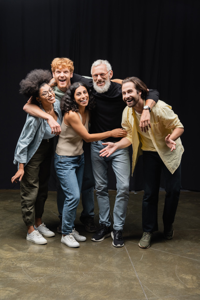young and joyful interracial actors looking at camera while embracing bearded producer in theater - Fotografie, Obrázek