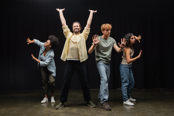 young man standing with raised hands and closed eyes near multiethnic students rehearsing on stage in theater - Fotoğraf, Görsel