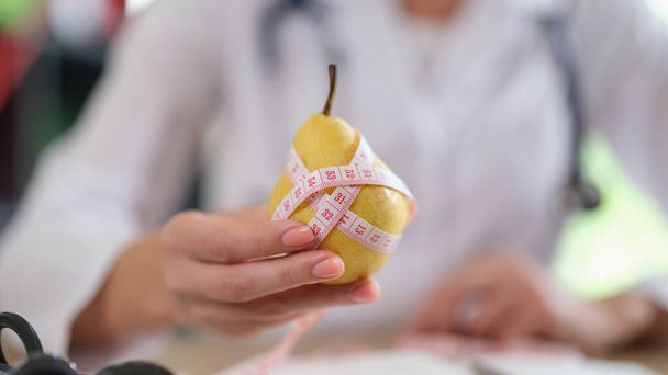 医者は果物や食べ物をビタミンで食べることを提案する。医務室に座っている間、測定テープで熟した黄色の梨を示す栄養士. - 写真・画像
