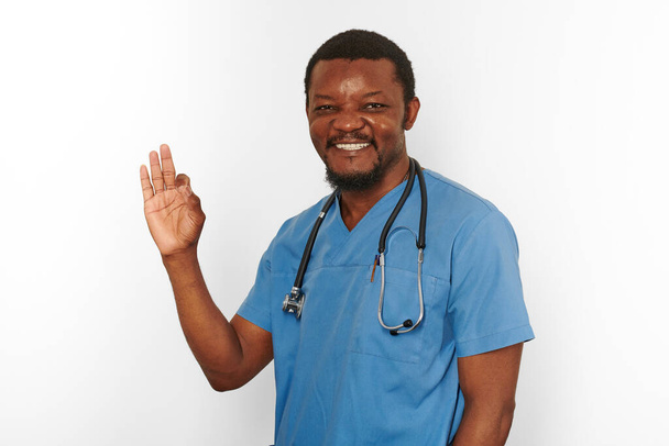 Smiling black surgeon doctor bearded man in blue coat with stethoscope makes OK gesture, isolated on white background. Happy adult black african american practicing surgeon portrait - Photo, Image