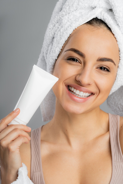 Smiling woman with towel on head holding cosmetic cream isolated on grey  - Foto, immagini