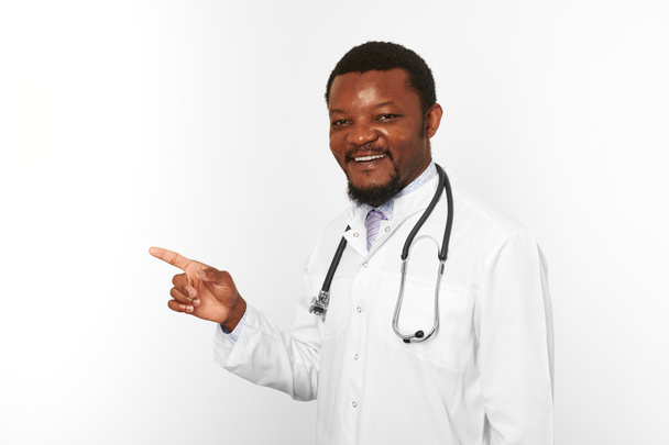Smiling black bearded doctor man in white coat with stethoscope points finger to left, isolated on white background. Adult black african american physician therapist portrait, candid male emotion - Photo, Image