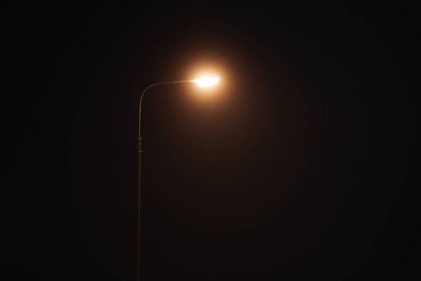 One night lamppost shines with faint mysterious yellow light through evening fog. Streetlight shine at quiet city night, magic atmospheric light in mystical darkness, copy space - Zdjęcie, obraz