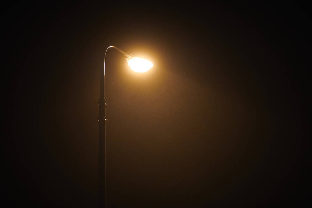 One night lamppost shines with faint mysterious yellow light through evening fog. Streetlight shine at quiet city night, magic atmospheric light in mystical darkness, copy space - Zdjęcie, obraz