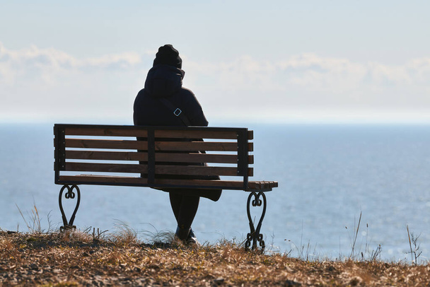 Single girl in a black jacket and hat sitting on bench at cliff at front of sea, peaceful and quiet place for thinking alone, loneliness and loss of loved one concept. Pacifying view of marine horizon - Foto, Imagem