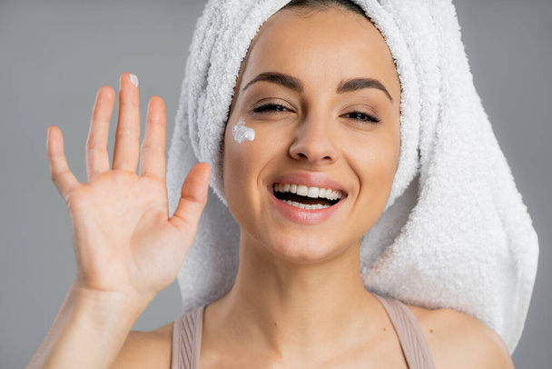 Cheerful woman with towel on head and cosmetic cream on cheek isolated on grey  - Foto, imagen