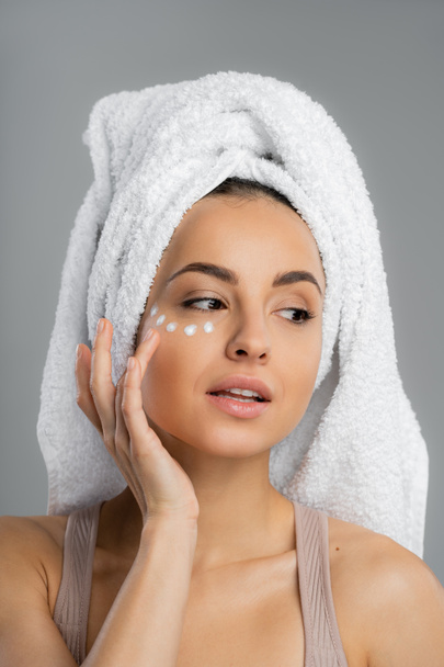 Young woman with towel on head applying cream under eye isolated on grey  - Foto, Imagen