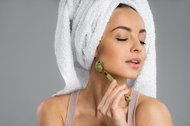Woman with towel on head using jade roller isolated on grey  - Foto, imagen