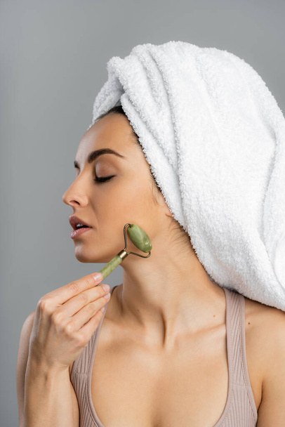 Pretty young woman with towel on head massaging face with jade roller isolated on grey  - Foto, immagini