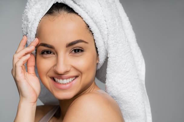 Portrait of smiling woman with towel on head touching face isolated on grey  - Foto, immagini