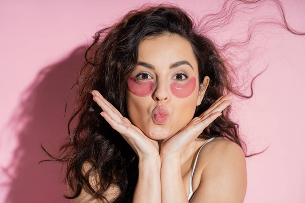 Curly woman with hydrogel eye patches pouting lips on pink background  - 写真・画像