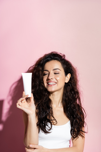 Cheerful freckled woman looking at cosmetic cream on pink background  - Photo, image