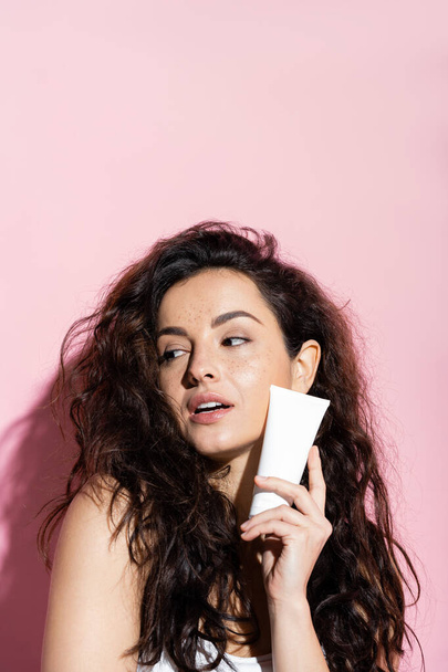Pretty woman with freckled skin holding cosmetic cream on pink background  - Foto, afbeelding