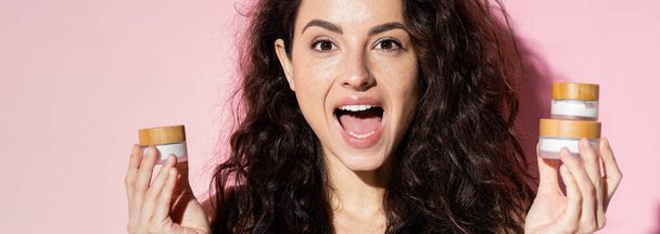 Excited freckled woman holding cosmetic creams on pink background, banner  - Φωτογραφία, εικόνα