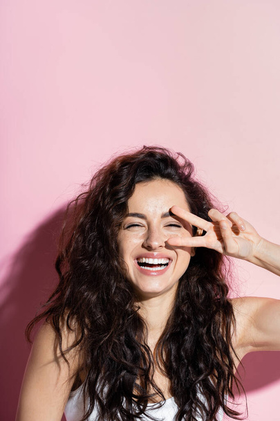 Cheerful curly woman showing victory sign on pink background  - Photo, Image