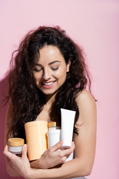 Positiivinen brunette nainen katselee kosmeettisia valmisteita vaaleanpunaisella taustalla  - Valokuva, kuva