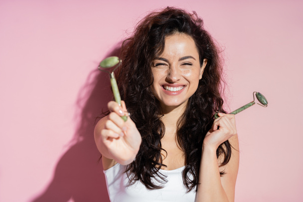 Positive freckled woman holding jade rollers on pink background  - Фото, изображение